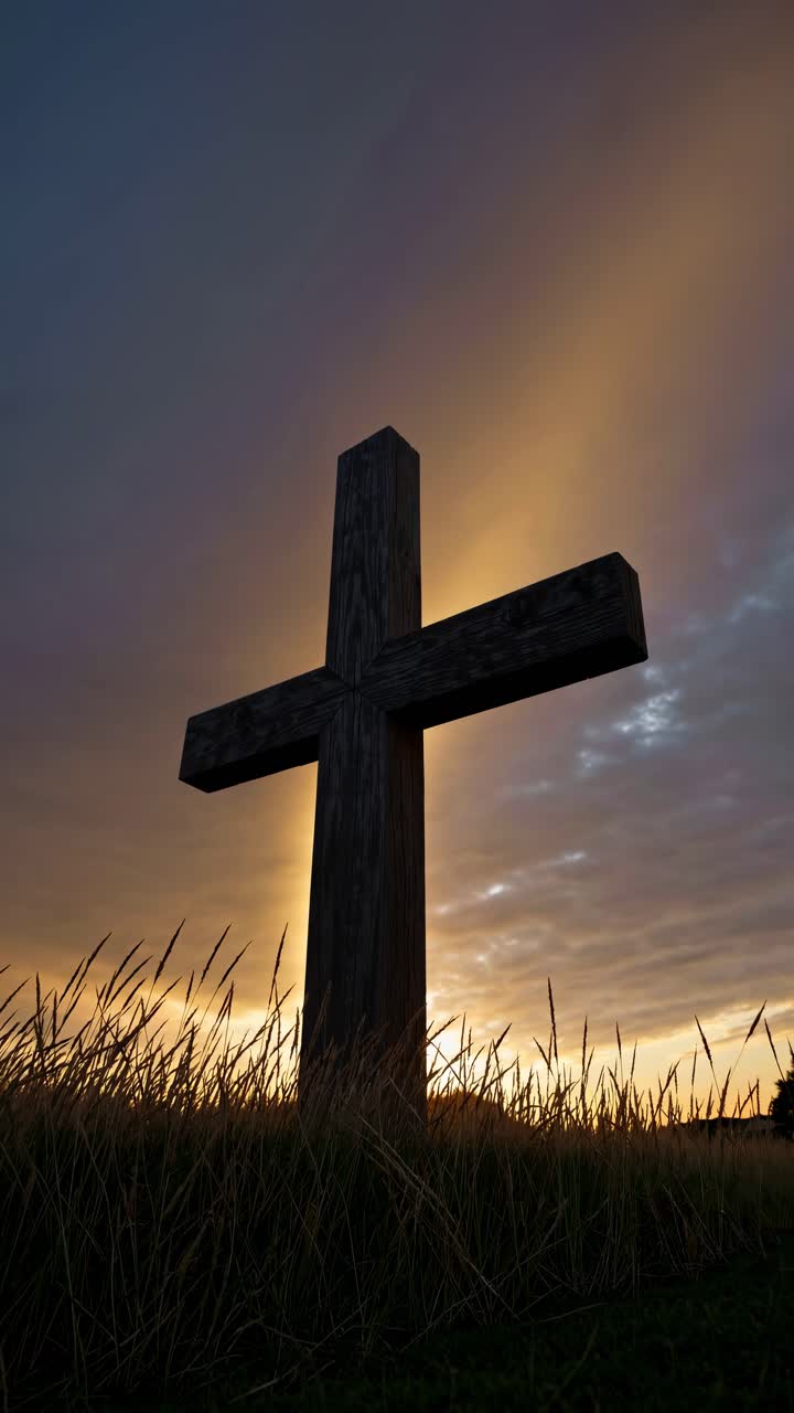 Low-angle video shot of a wooden cross silhouetted against a dramatic sunset sky, surrounded by tall