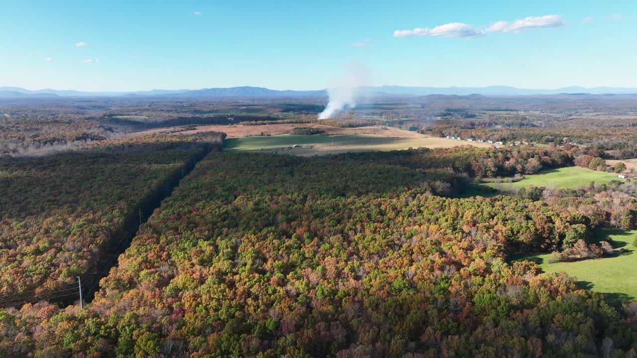 Colorful american countryside with forest trees and burning fire on field in background. Sunny day in autumn season. Smoke rising into sky. Drone forward wide shot.