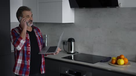 modern aged man using laptop in kitchen and talking by phone at the same time