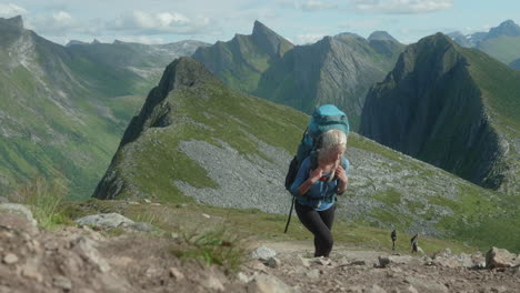 Pretty-Blonde-Female-Hiker-Walking-Up-a-Steep-Hill-infront-of-an-Impressive-Backdrop-of-Mountains-and-Fjords,-on-a-Sunny-Summer-Day-in-Northern-Norway,-Senja