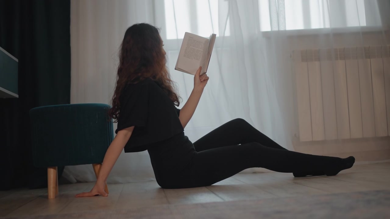 Donna seduta sul pavimento in moquette accanto al divano, appoggiata all'indietro su una mano mentre tiene un libro aperto vicino al viso sotto la delicata luce della finestra, creando una tranquilla e accogliente scena di lettura casalinga con una postura rilassata.