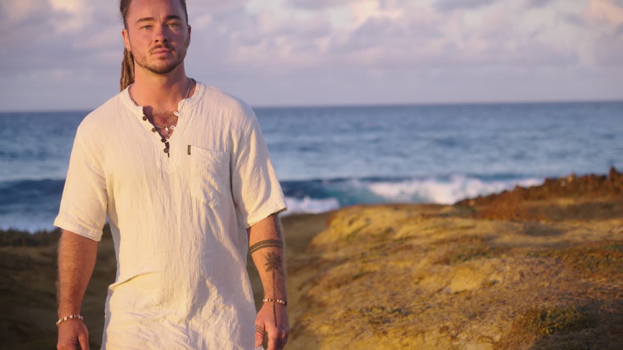 A man in white clothing walks confidently along a coastal path, surrounded by rocky terrain and the blue ocean horizon under soft morning light