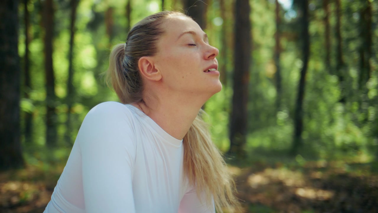 Young woman rests in sunlit forest, breathing deeply. She wears white top and pink pants, hair tied back. Eyes closed, calm expression shows peacefulness. Forest floor covered in soft moss