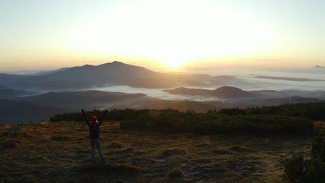 a hiker standing on a rock and raising hands in a gesture of victory on top of a mountain during sunrise