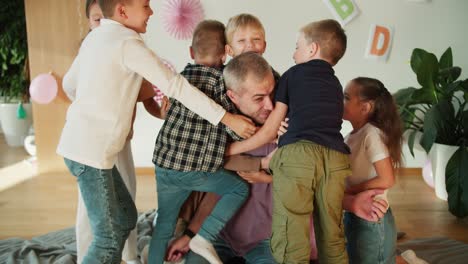 A-group-of-children-hug-their-teacher,-a-man-with-gray-hair-in-a-purple-T-shirt,-at-their-first-lesson-in-preparing-children-for-school-in-a-special-club.-The-happy-children-love-their-teacher-and-hug-him