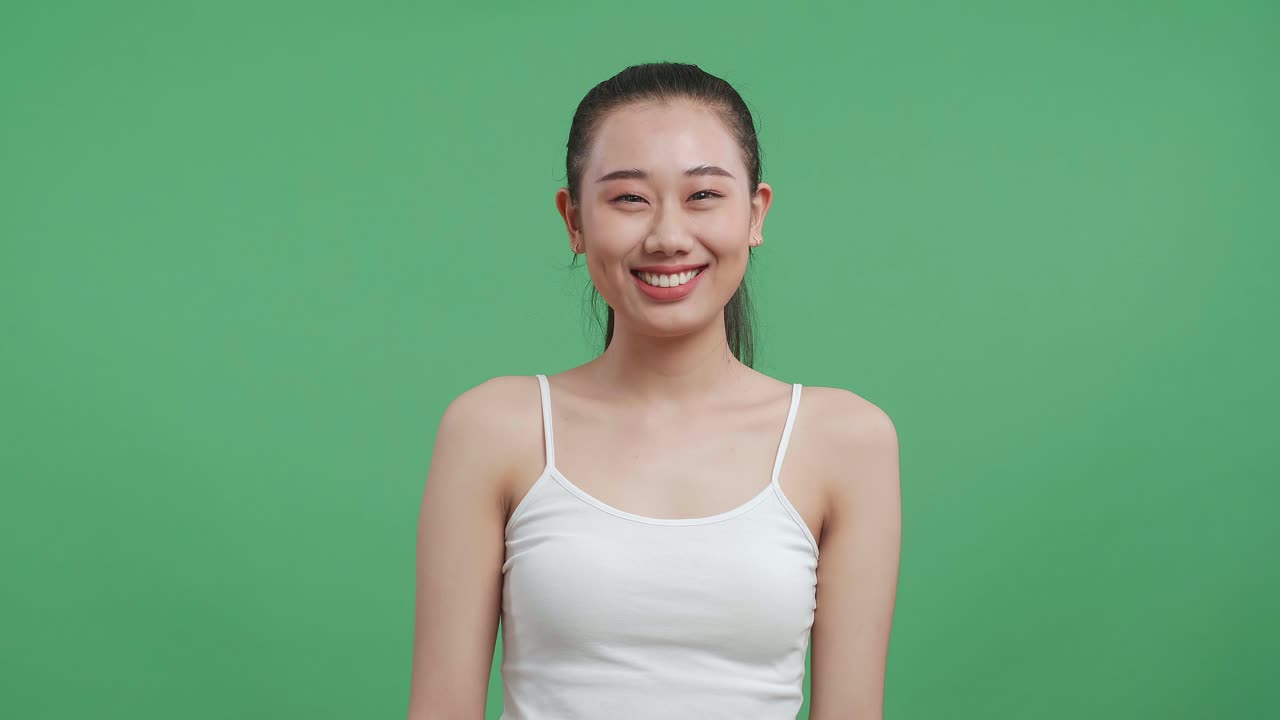 백색 카미솔을 입은 아시아 여성이 스튜디오의 녹색 스크린 배경에서 따뜻하게 미소 짓고 있습니다.