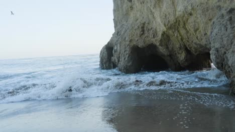 waves gently crash against a rocky cave along a serene beach, with reflections forming on the wet sand under soft daylight