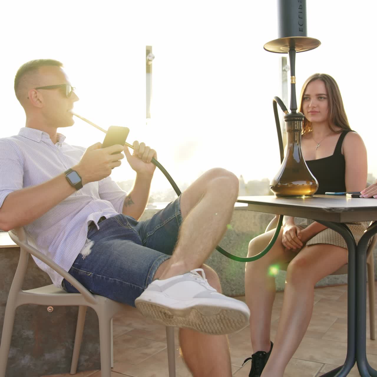 Confident relaxed man in sunglasses smoking hookah and looking at his phone. Girls sitting next enjoying city view from the roof top. Setting sun backdrop. Low angle view