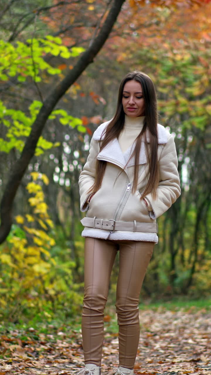 Relaxed long-haired brunette tossing fallen leaves walking by the road. Calming leisurely stride in the autumn park. Vertical video
