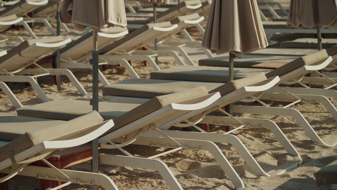 Tomme strandliggestole med parasoller på en sandstrand, der venter på turister, der skal slappe af og nyde sommerferien.