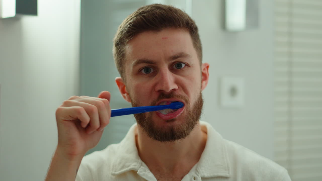 A man stands by the mirror brushing his teeth as part of his morning routine, illuminated by soft bathroom light. A clean and relatable daily lifestyle moment