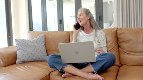 mujer caucásica madura sentada en el sofá, hablando por teléfono, sosteniendo una computadora portátil