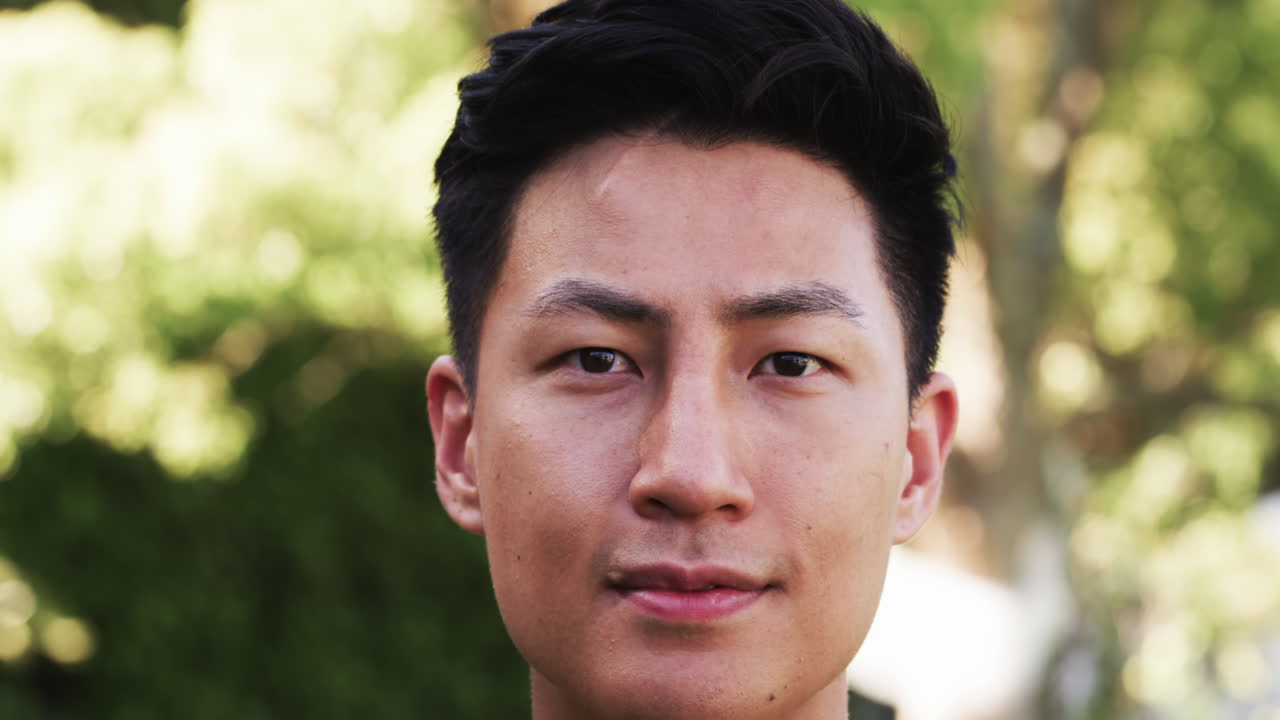 Confident Asian man outdoors, looking at camera with serene expression, in park
