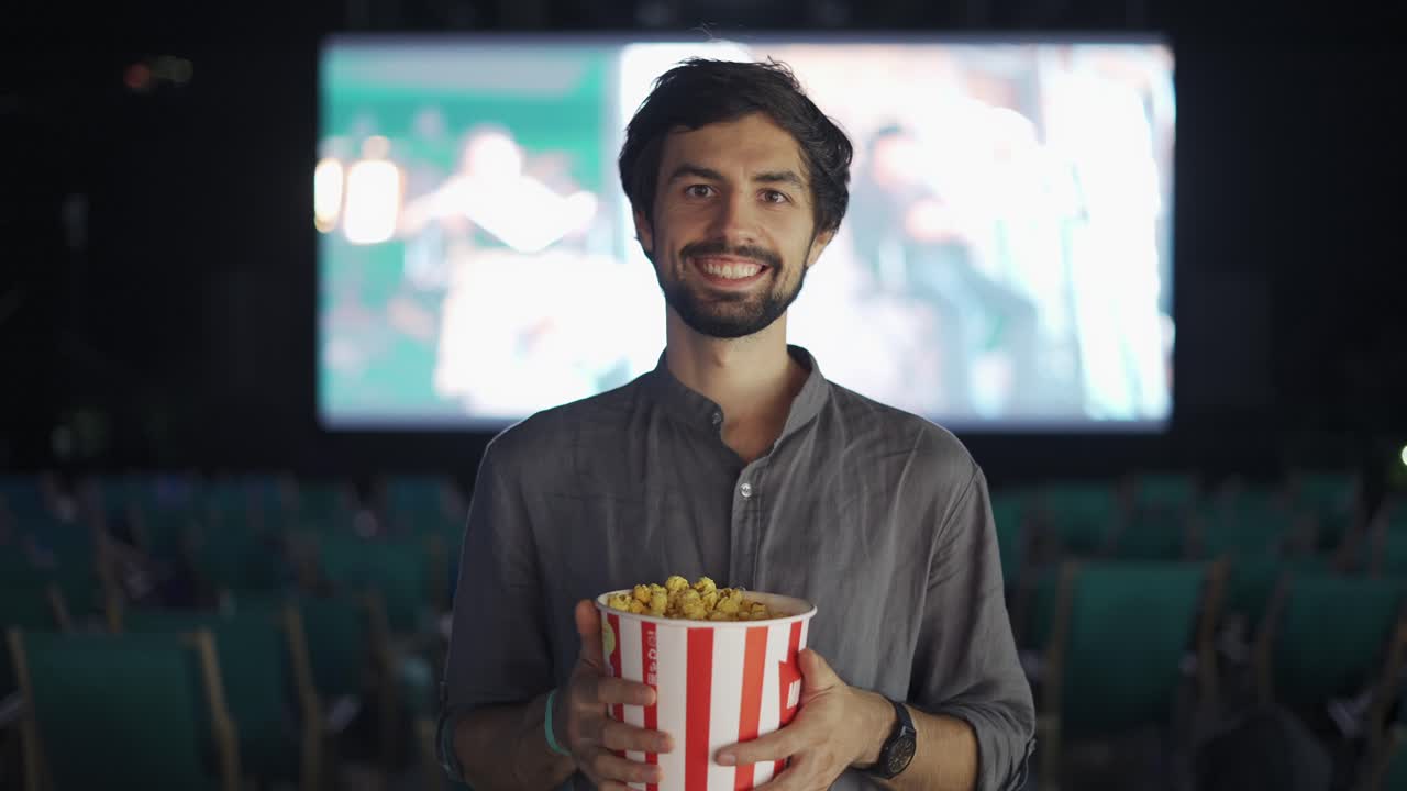 A bearded guy standing with bucket of popcorn at the cinema, smiling to the camera