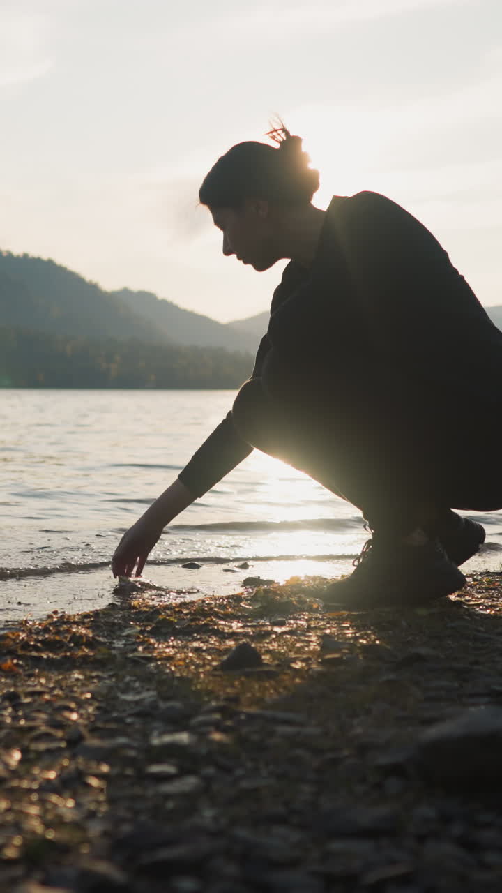Lady squats stroking water of lake at sunset. Woman searches inner harmony during solo journey and dreaming about future. Calm atmosphere at twilight enhances experience