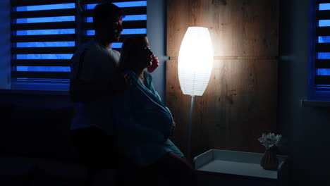 future parents in the dark hospital room with a nightlight. man soothes his wife holding her from the back.