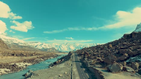 drone flies slowly upwards next to a road and a river in skardu with a view of a high mountain range covered in snow