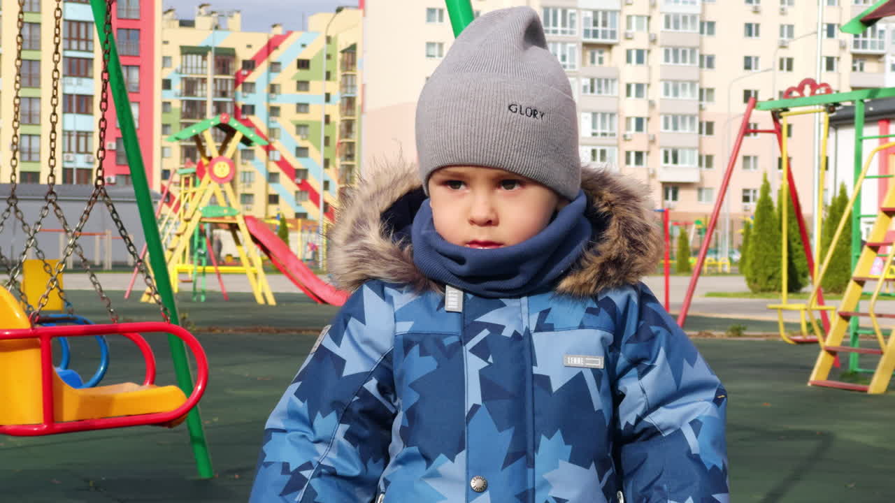 Serious Caucasian toddler in grey cap and warm jacket stands looking around. Little kid on the playground in autumn.