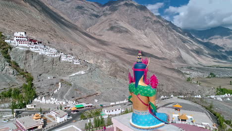 Vista-Aérea-De-Una-Estatua-Alta-De-Lord-Budha-Con-Un-Antiguo-Monasterio-En-El-Fondo-Situado-Sobre-Una-Montaña-A-Distancia