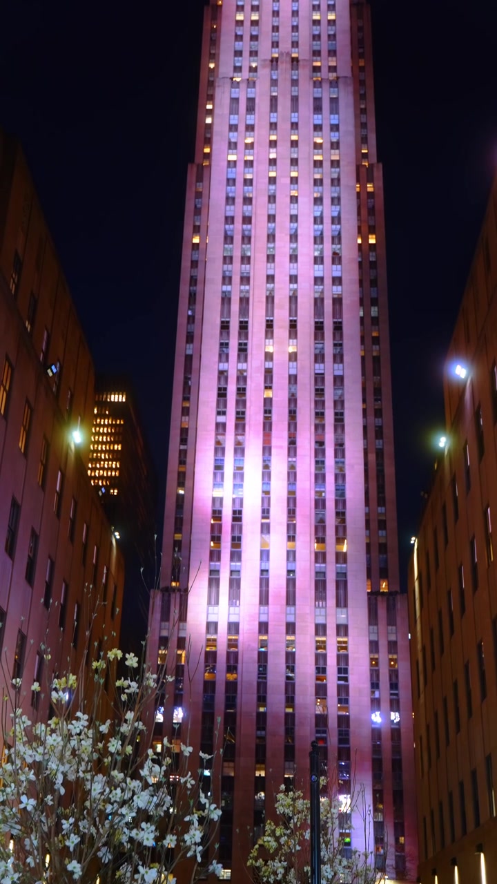 Arranha-céu Art Déco iluminado em rosa à noite. Perspectiva vertical de fachada de calcário com janelas iluminadas ladeada por edifícios vizinhos.