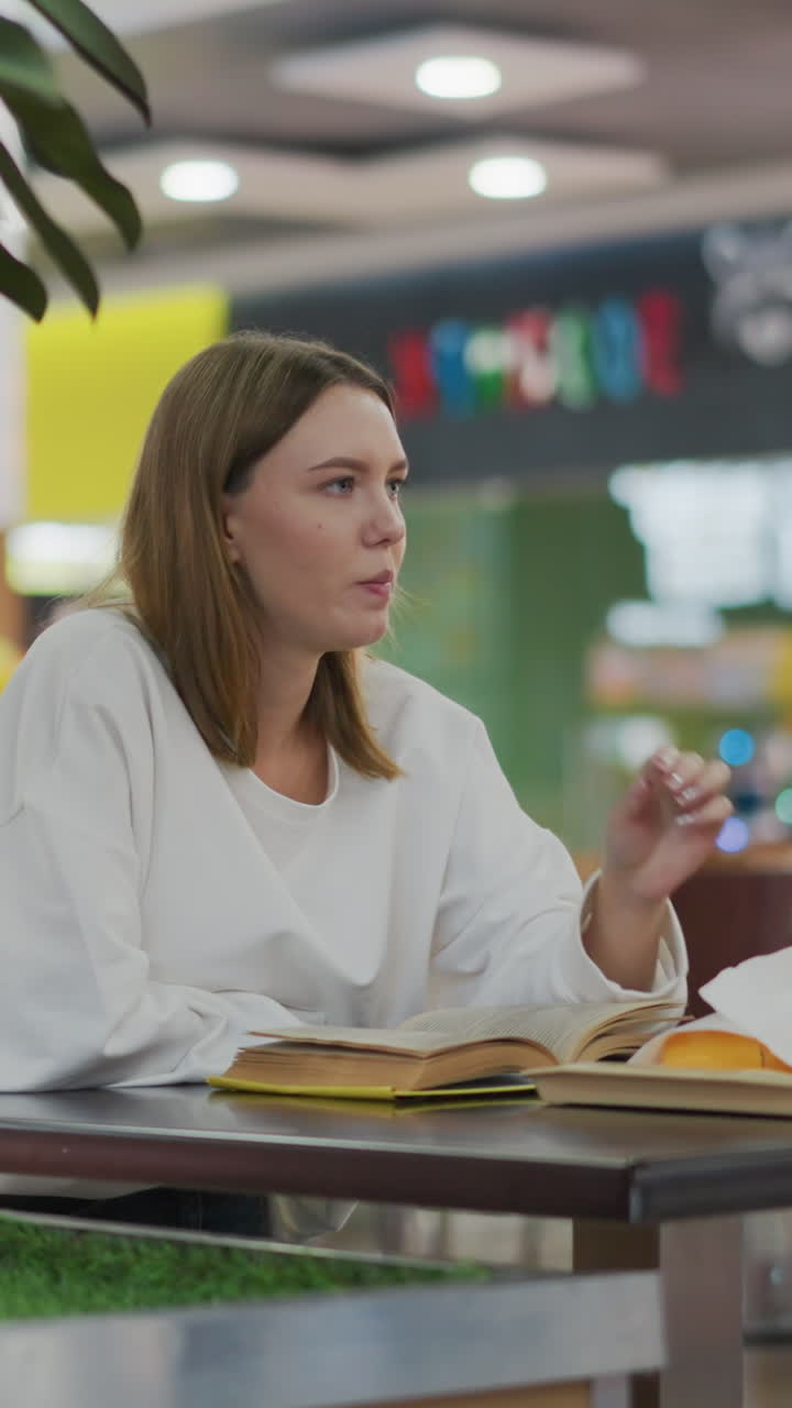 primer plano de una mujer comiendo un bocadillo mientras lee un libro en un restaurante moderno, fondo vibrante con iluminación suave, bebida en la mesa y bolsas de compras a la vista