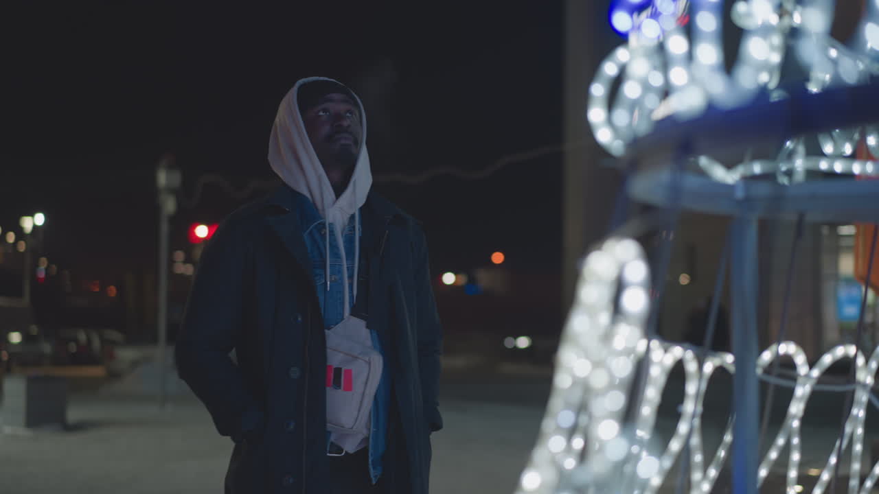 Man standing outdoors at night, gazing up at glowing festive lights and illuminated number-shaped decoration in front of a shopping complex. Winter atmosphere, city lights, and holiday spirit