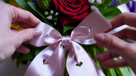 hands tying a pink ribbon bow with hearts on a red rose bouquet