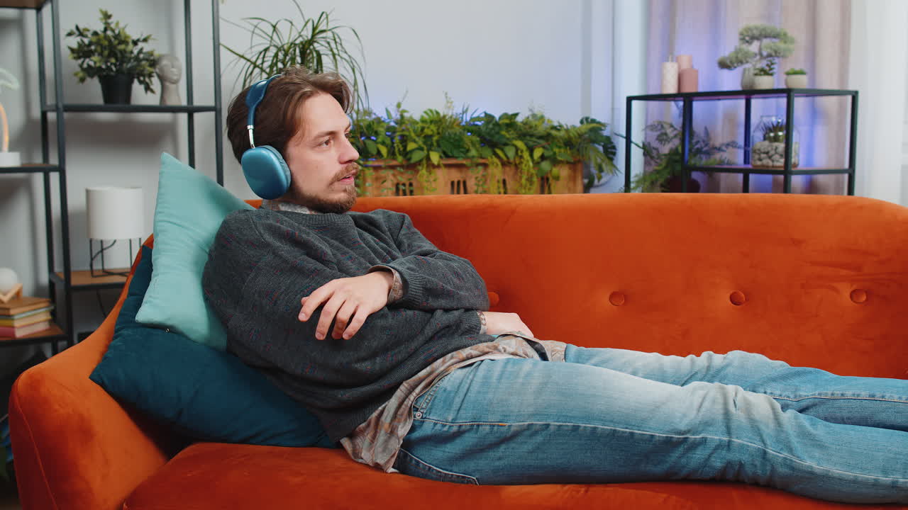 무선 헤드폰을 착용한 행복한 남자가 가정 소파에 누워서 좋아하는 록 앤 롤 음악을 듣고 휴식을 취하고 있습니다.