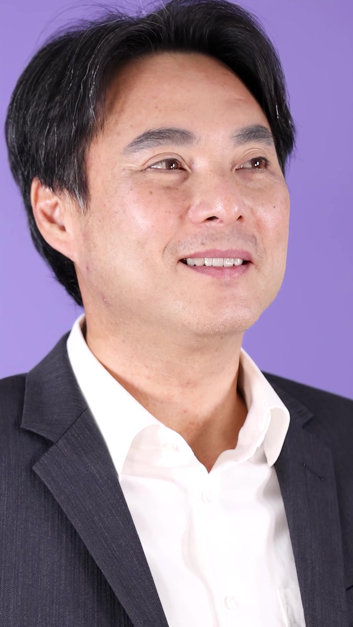 Uomo asiatico in abito sorride con sicurezza, studio ben illuminato, telecamera fissa, sfondo viola pulito