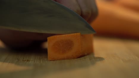 toma de primer plano fija de verdura de zanahoria de forma cuadrada pelada en la mano del chef mientras la corta y la corta en rebanadas usando un cuchillo de cocina en una tabla de cortar de madera, 4k