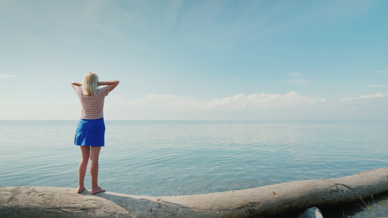 한 젊은 여성이 통나무 위에 서서 하늘이 바다와 합쳐지는 아름다운 풍경을 감상한다