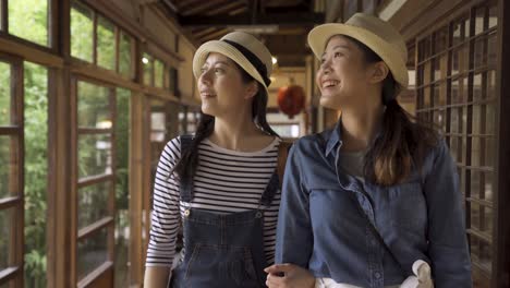 two young girls tourists visiting historic japanese traditional house walking in hallway discussing talking. asian female best friends relax chatting standing in corridor wooden building point finger