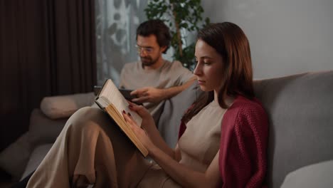 Side-view-of-a-young-brunette-girl-in-a-red-sweater-reading-a-book-while-sitting-on-a-gray-sofa-with-her-boyfriend-in-glasses-who-is-looking-at-a-tablet-in-a-modern-apartment