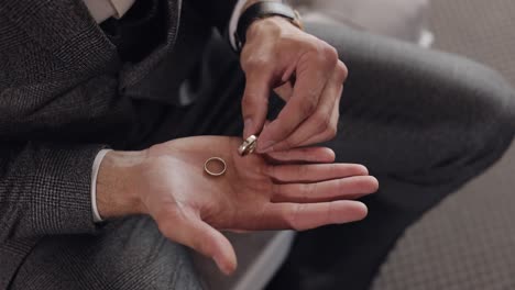 Groom-in-gray-jacket-holding-wedding-rings-on-palm-of-his-hand,-man-touches-the-rings,-slow-motion