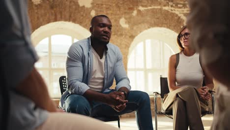 A-Black-man-in-a-blue-shirt-expresses-his-thoughts-during-group-therapy-in-a-brick-room-with-yellow-lighting.-A-girl-in-a-white