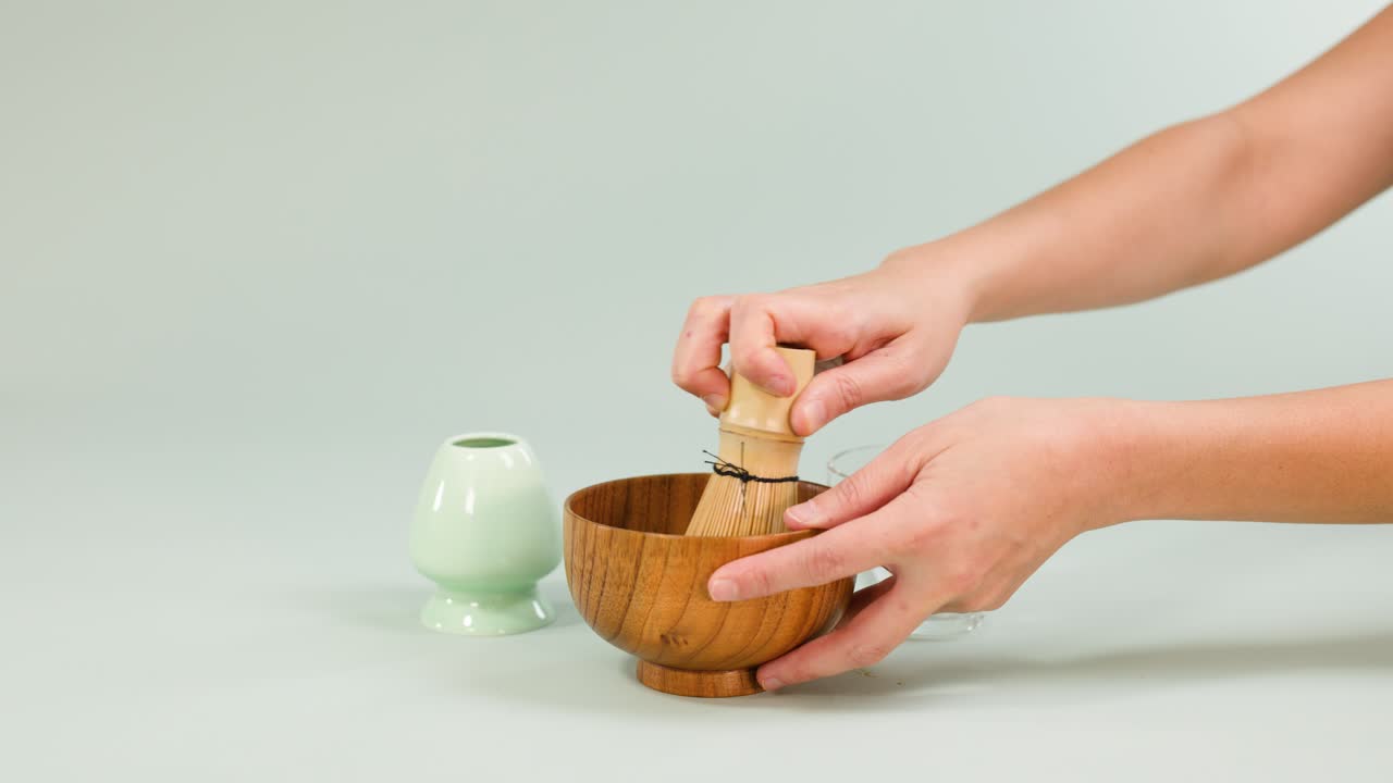 Hands whisk matcha in a wooden bowl using bamboo whisk, minimal clean background, soft lighting