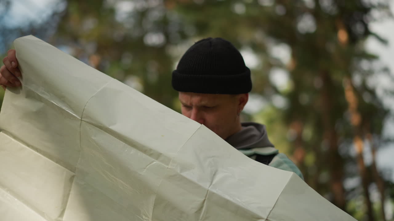 white hiker reads large map in forest, unfolding paper among pine trees, scanning contours and compass, plotting route and checking direction, introspective scout smoothing creased sheet