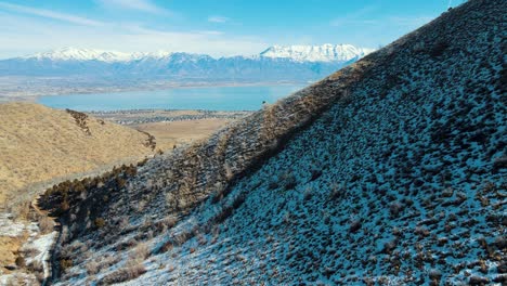 A-snowy-hillside-looking-down-a-canyon-to-a-valley-with-a-lake-and-snow-capped-mountains---aerial-parallax-view