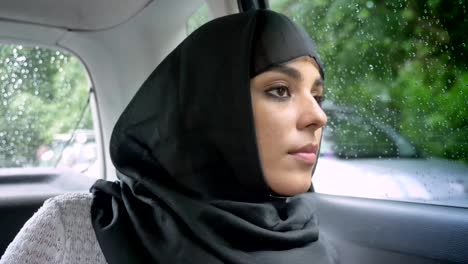 young muslim woman in hijab is looking out for location in rainy window in car, transport concept, weather concept