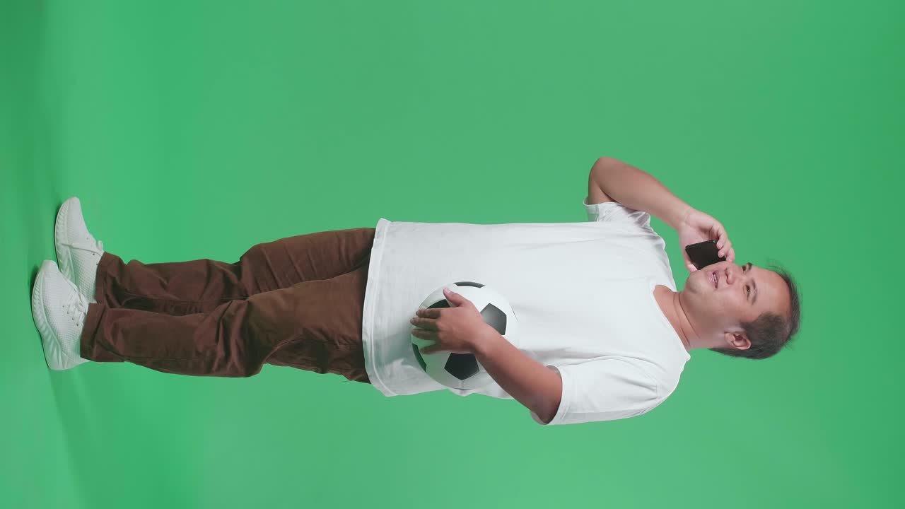 녹색 화면 배경에서 축구를 응원하는 동안 스마트폰으로 말하는 공을 가진 아시아 남자의 몸이 가득 차 있습니다.