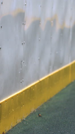 el sutil reflejo de una persona en una pared metálica con el impacto dinámico de una pelota de fútbol capturado en medio del movimiento, enfatizado por el borde amarillo de la pared y el juego de sombras, creando energía bajo la brillante iluminación nocturna
