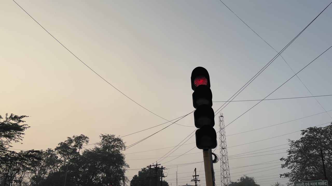 Foto estática em ângulo baixo de um semáforo vermelho brilhando em um cruzamento, silhuetado contra o céu noturno com linhas de energia e árvores formando um plano de fundo urbano