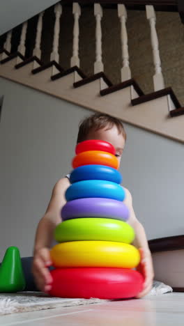 lovely smiling baby boy sits on the floor building a pyramid cheerfully. low angle view on the toddler playing at home. vertical video.