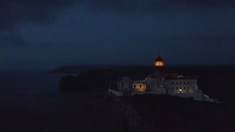 Static-aerial-view-of-Lagos-lighthouse-against-cloudy-dark-blue-sky-at-dusk,-light-flashing-creates-lens-flare