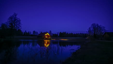 Tiro-Estático-Desde-La-Noche-Hasta-El-Movimiento-De-Estrellas-Del-Cielo-Nocturno-En-Timelapse-En-El-Campo-Rural-Con-La-Vista-De-Un-Lago-Frente-A-Una-Pequeña-Cabaña