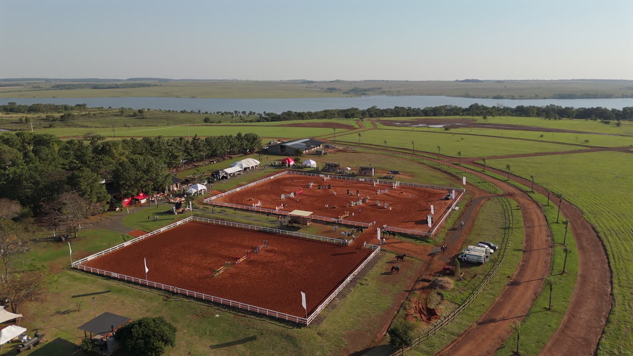 Drone captures orbiting aerial view of equestrian training complex with two fenced red dirt arenas, curved dirt tracks, tents, and fields under open sky, rural sports facility in wide daylight motion.