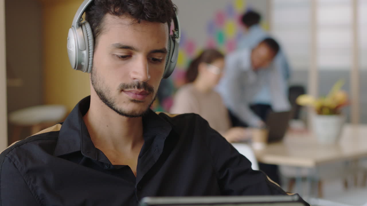joven hispano usando una tableta digital, un ordenador, dibujando, navegando en línea, compartiendo mensajes en las redes sociales, estudiantes dibujando, disfrutando escuchando música, comprobando el smartphone en una oficina moderna