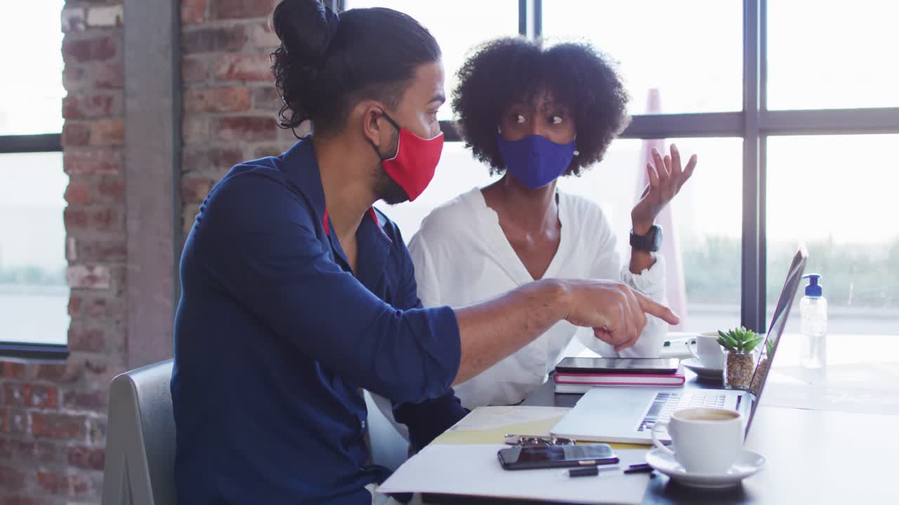 Diverse couple wearing face masks sitting in cafe using laptops and talking