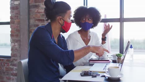 Diverse-couple-wearing-face-masks-sitting-in-cafe-using-laptops-and-talking