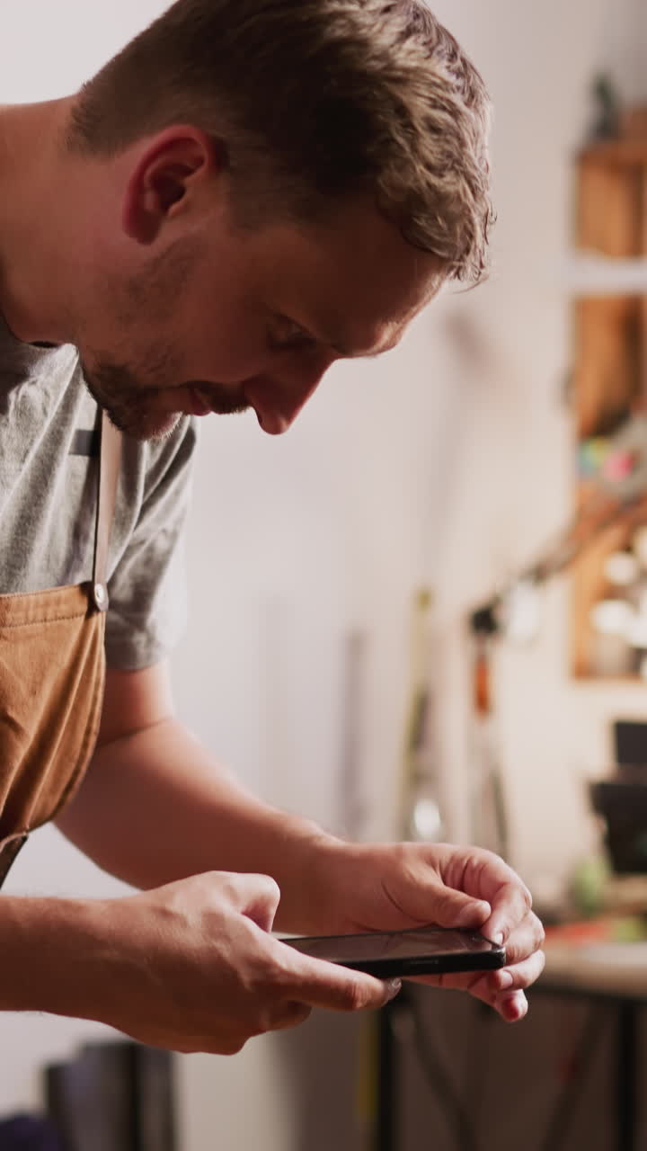 Leathersmith in apron takes photo of handmade leather wallet on wooden tray with mobile phone in design studio closeup. Craftsman shows product in social media